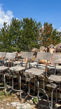 A row of old weathered wooden bar chairs with rusty metal frames outdoors against a backdrop of green trees and a clear blue sky.