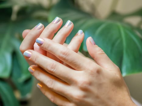 Female Hands With Light Purple Chrome Ombre Nails Manicure. Woman Fingernails With Pink And Blue Gradient Aura Nails On Green Lush Background. Aesthetic Visual Of Trendy Manicure With Nail Art.