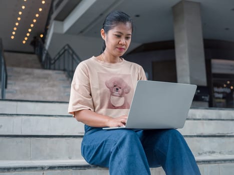 Asian business woman with laptop computer working outside looking at screens in the business district, Professional woman sitting outdoors, A woman works on a computer on an outdoor staircase.