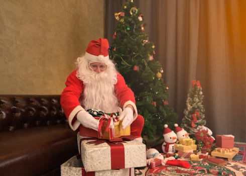Santa Claus in red suit sitting on couch in living room at home next to decorated Christmas tree, snowman decorations and presents wrapping gift boxes around. Christmas and New Year Holiday celebration concept