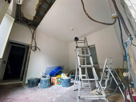 A room in the middle of a renovation process, featuring a ladder, exposed electrical wires, construction materials, and tools scattered on the floor.