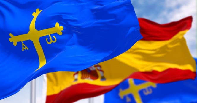 The Asturias flags waving with Spanish flag on a clear day. Autonomous community in northern Spain. Blue with golden victory cross. 3d illustration render. Rippling fabric. Selective focus