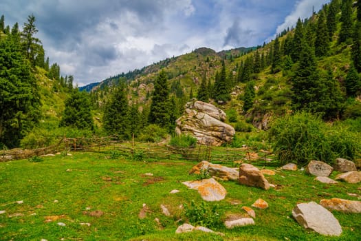 Green mountain landscape with rural stick fence and large rock at summer day in Kyrgyzstan
