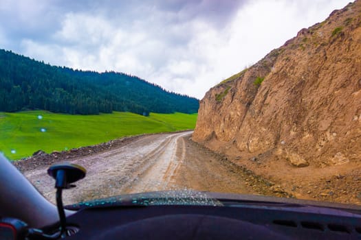 driving along the dirt road at rainy summer day in mountains with spruce forest on mountain slope, view from driver perspective