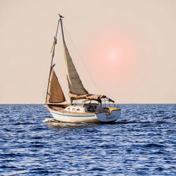 A sailboat glides smoothly over tranquil waters as the sun sets on the horizon, creating a serene and picturesque atmosphere. The gentle breeze fills the sails, enhancing the peaceful ambiance.