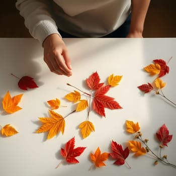 A person carefully arranges vibrant red, orange, and yellow autumn leaves on a bright white table. The warm sunlight illuminates the scene, highlighting the fall colors.