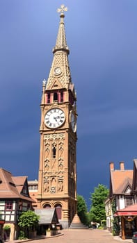 A tall, ornate clock tower stands proudly against a dramatic blue sky, surrounded by charming, traditional buildings, creating a picturesque view filled with historical charm.