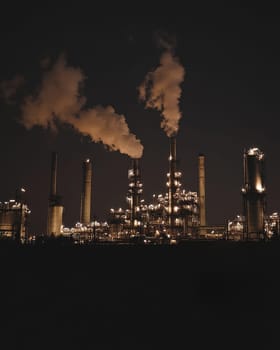 Illuminated industrial plant with towering smokestacks emitting steam against a dark night sky.