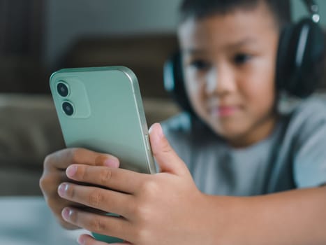 Cute elementary school boy wearing headphones holding a smartphone. Boy studying online on smartphone or homeschooling, listening to music or playing games, Concept of online learning via smartphone.