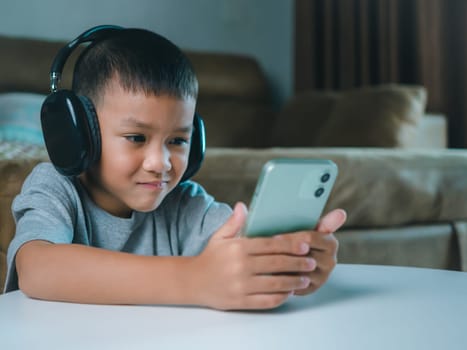 Cute elementary school boy wearing headphones holding a smartphone. Boy studying online on smartphone or homeschooling, listening to music or playing games, Concept of online learning via smartphone.