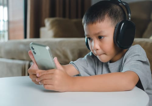 Cute elementary school boy wearing headphones holding a smartphone. Boy studying online on smartphone or homeschooling, listening to music or playing games, Concept of online learning via smartphone.