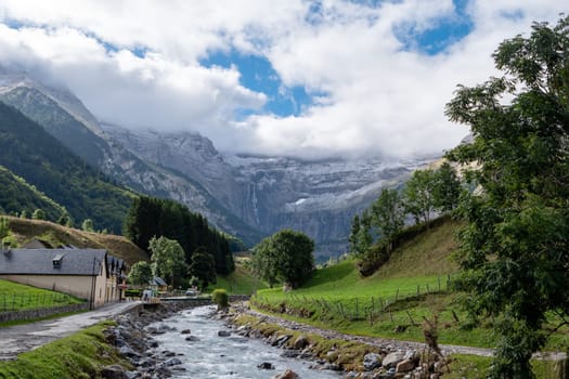 The Cirque of Gavarnie, french Pyrenes mountains. The mountains are covered in snow and the sky is cloudy. The river is surrounded by a lush green valley