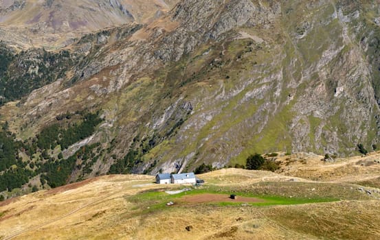 A small house sits on a hillside with a view of the mountains. The house is surrounded by a lush green field. The scene is peaceful and serene