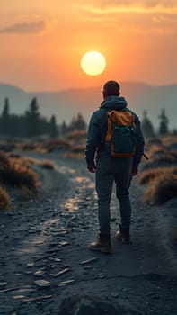 A lone hiker stands on a mountainous path, gazing at the vibrant sunset. The golden sun casts a warm glow, illuminating the serene landscape filled with distant hills and rocky terrain.