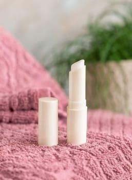 Opened lip balm stick on pink folded towel near green plant in bathroom,p, closeup, mockup. Lifestile scene with beauty product, everyday skincare routine