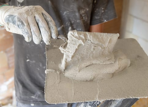 Craftsman in dirty gloves puts plaster, mortar or cement on a metal trowel. Men in a gray shirt with visible dust and plaster on clothing during construction or renovation process