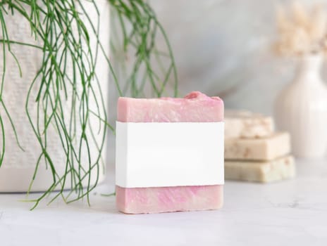 Pink soap bar with blank label on white marble countertop near potted green plant in bathroom, close up, packaging mockup. Natural cosmetic product for face or body care