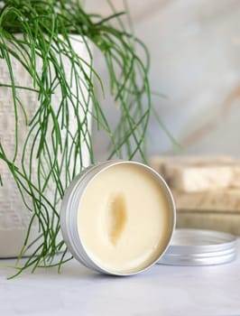 Opened body butter jar on white marble countertop near potted green plant in bathroom, close up. Natural cosmetic product for face or body care, everyday lip care routine