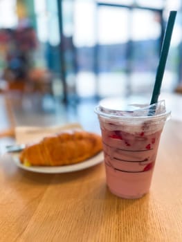 Refreshing pink drink in a plastic cup sits on a wooden table in a cafe. And delicious croissant on a plate, cozy and inviting atmosphere