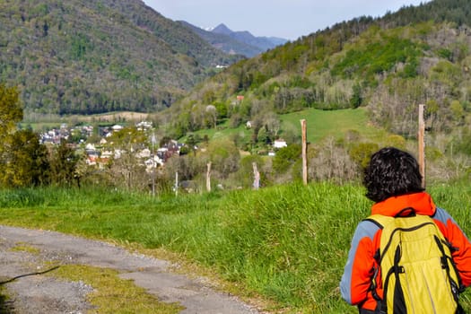 A woman is walking down a dirt road with a yellow backpack. The road is surrounded by mountains and the sky is clear. The woman is enjoying the scenery and the peacefulness of the area