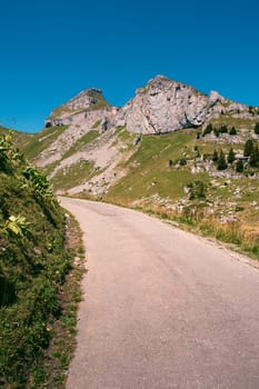 Concrete mountain road between alpine meadows and rocks, peaks of Tour de Mayen and Tour d'Ai in the background on a bright summer day. Leysin, Vaud, Switzerland