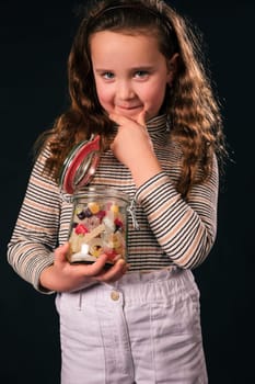 8 Year old girl holds glass jar with colorful candy holding hands on chin pondering and looking at camera. eat or not eat. dark background