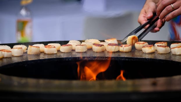 Delicious scallops being flipped over grilling on round steel iron firepit hearth table surface brazier barbecue, shot with shallow dept of field