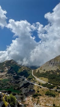 A scenic view of a curving mountain road winding through green hills under a sky filled with dramatic clouds. The road leads through a rugged landscape.
