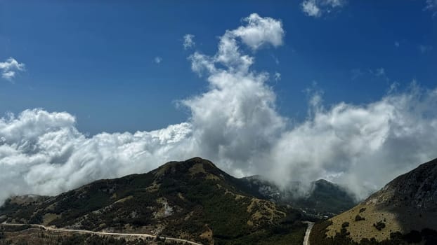 A captivating view of clouds rolling over a lush mountain range, with a winding road cutting through the green hills, under a bright blue sky.