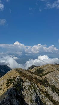 A wide view of a rugged mountain ridge, with rocky outcrops and sparse vegetation, set under a vast, cloud-filled blue sky.