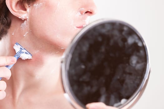 Young man carefully shaving his face with a blue razor in a bathroom mirror, focusing on grooming routine