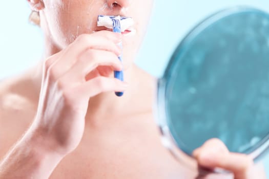 Young man shaving in a blue bathroom, carefully removing hair to reveal smooth skin. His daily grooming routine ensures healthy skin