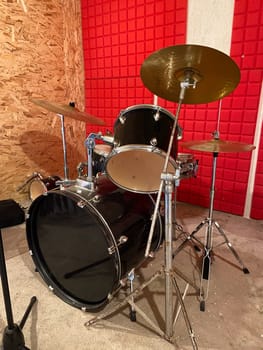 Classic black drum set is positioned in a studio with vibrant red soundproof walls. Shiny black drums and bright red background creates a dynamic and energetic music atmosphere. Musical instrument and creative art concept.