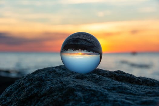 Glass ball on a rock at sunset on the beach, the sea and the setting sun are reflected in the ball