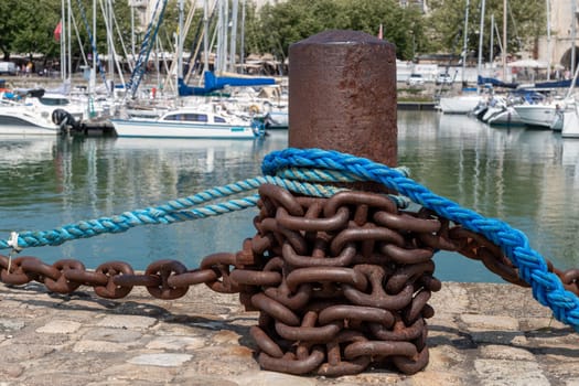 A chain is wrapped around a metal post with a blue and white rope. The post is in front of a marina with several boats docked