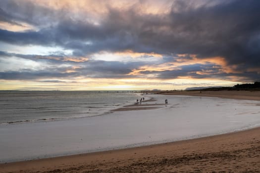 The beach is a serene and tranquil place, with a few people walking along the shoreline. The sky above is cloudy, creating a moody atmosphere. The water is calm, and the sand is soft and inviting