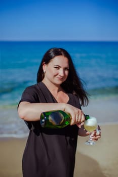 A brunette girl in a black dress pours a glass of champagne on the seashore