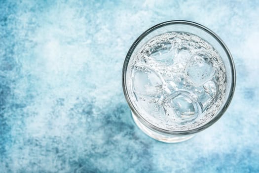 A glass filled with sparkling water and ice cubes sits on a textured blue surface reflecting light and creating bubbles.
