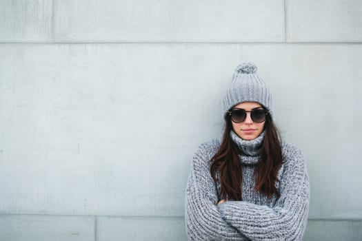 A young woman stands against a textured wall wearing warm gray knitwear and sunglasses showcasing a stylish winter look.