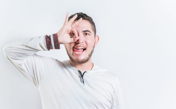 Funny young man making OK gesture looking through fingers isolated. Happy guy making ok gesture looking through fingers