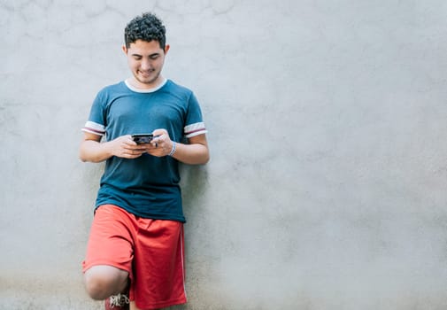 Happy guy leaning on a wall using smartphone. Smiling young man leaning on a wall sending a text message