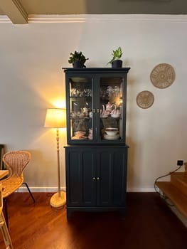 Classic wooden cabinet stands elegantly in a cozy room, displaying exquisite china and adorned with potted plants. warm lighting from a floor lamp adds a welcoming ambiance and highlights the intricate details of decor.