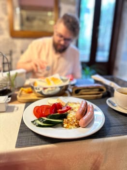 hearty breakfast is beautifully arranged on a plate, featuring sausages, fried eggs, and fresh vegetables. in the background, a person prepares to enjoy this appetizing meal, highlighting a cozy dining.