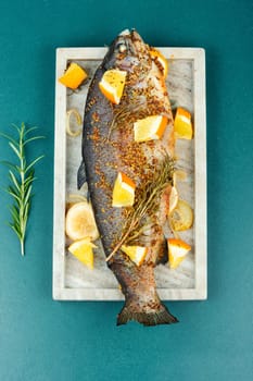 Whole baked trout on a stylish stone plate with orange on wooden background.