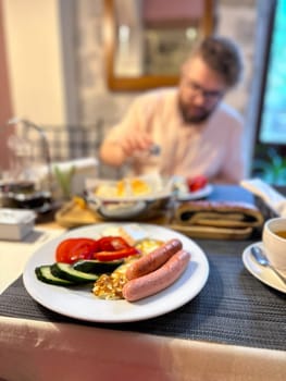hearty breakfast is beautifully arranged on a plate, featuring sausages, fried eggs, and fresh vegetables. in the background, a person prepares to enjoy this appetizing meal, highlighting a cozy dining.