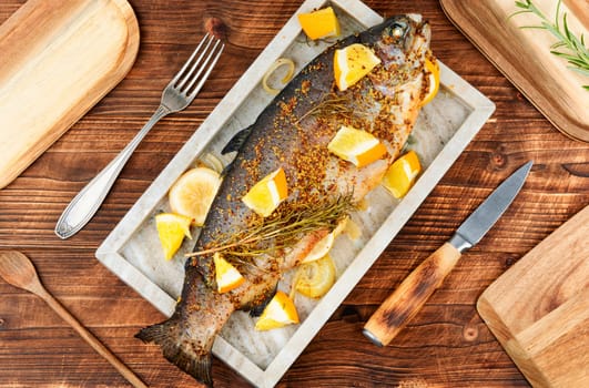 Whole baked trout on a stylish stone plate with orange on wooden background.