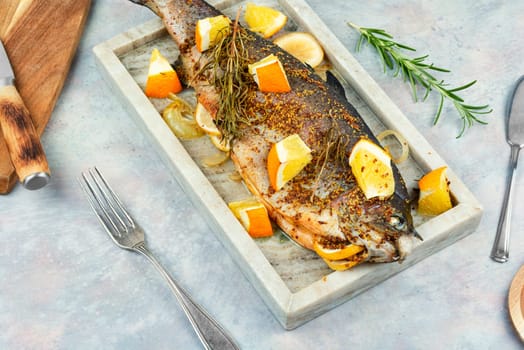 Whole baked trout on a stylish stone plate with orange on wooden background.