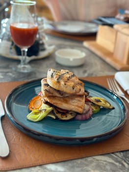 Grilled chicken stacked elegantly on a bed of vegetables, elegantly presented on a blue plate with a glass of juice nearby in restaurant. Culinary scene captures the artistry and appeal of modern dining in a beautifully set table environment