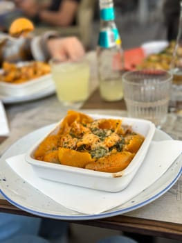Plate of savory pasta shells with creamy sauce is perfectly presented on white dish, depicting an inviting and appetizing meal in casual dining setting. the background hints at relaxed social gathering, enhanced by beverages and cozy atmosphere.