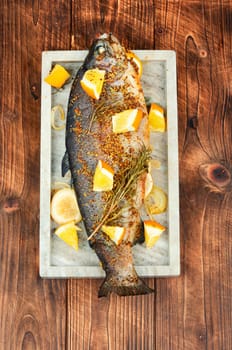 Whole baked trout on a stylish stone plate with orange on wooden background.